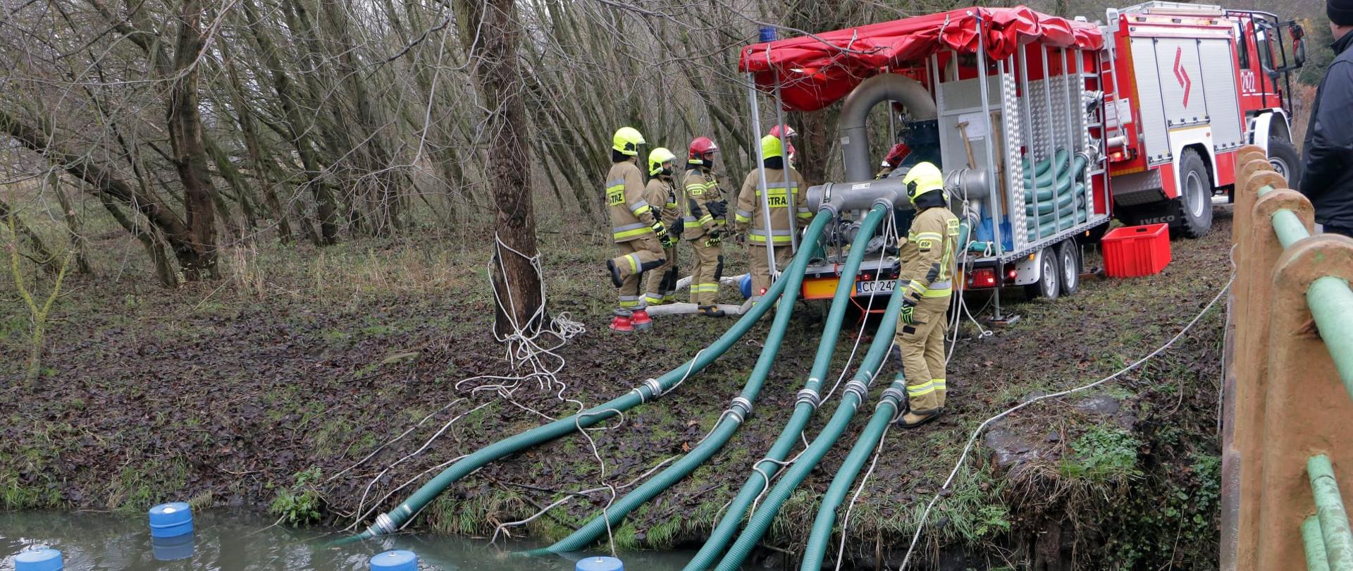 Zdjęcie przedstawia ciężarowe, biało-czerwono-srebrny samochód strażacki z przyczepą stojący przy rzeczce w lesie. Do przyczepy podłączone są zielone węże, które zanurzone są w w wodzie. Obok sprzętu pracują strażacy ubrani w piaskowe ubrania bojowe i hełmy.