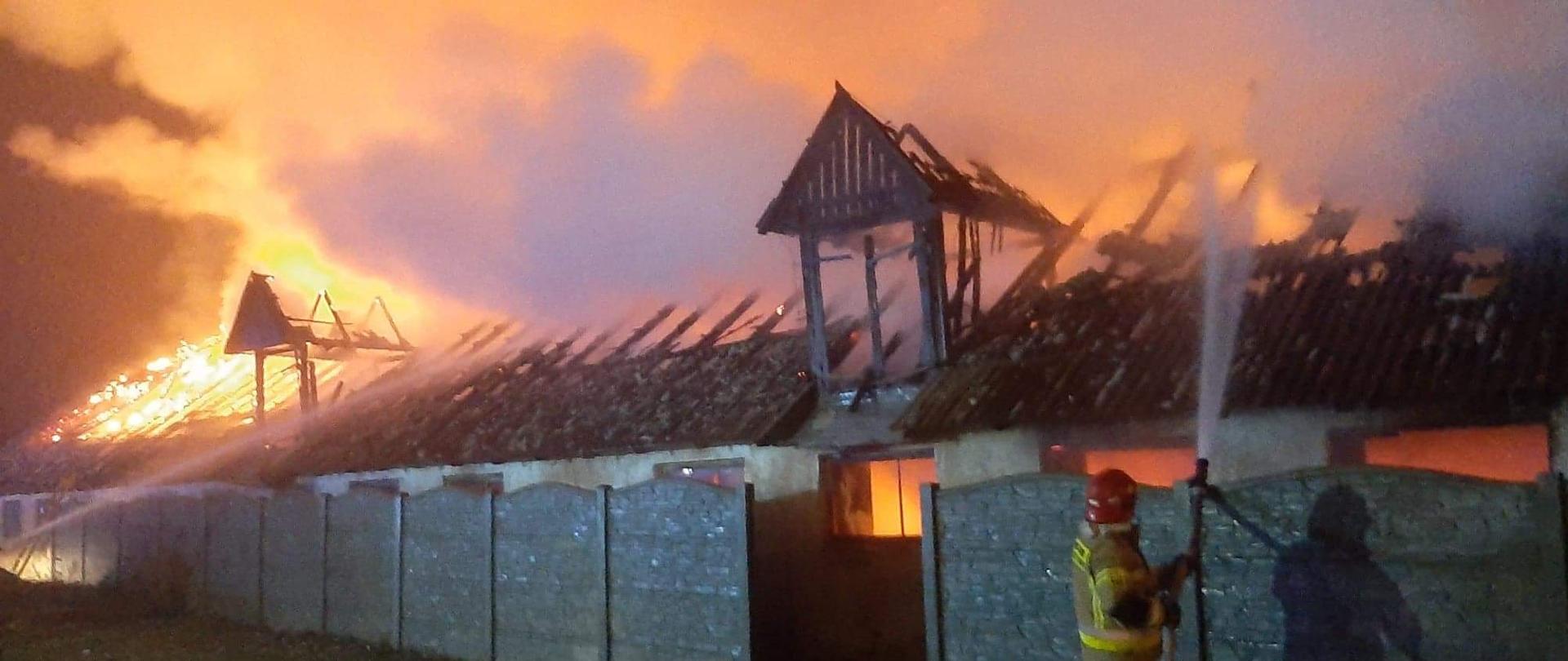 Na zdjęciu znajduje się budynek inwentarski, cały w ogniu, który znajduje się za betonowym ogrodzeniem. Po prawej stronie na zdjęciu przed ogrodzeniem stoi strażak i podaje prąd wody na ogień. 