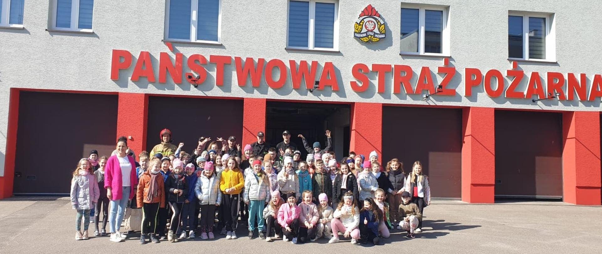 Uczniowie i nauczyciele ze szkoły stoją ze strażakami na placu przed budynkiem Komendy.