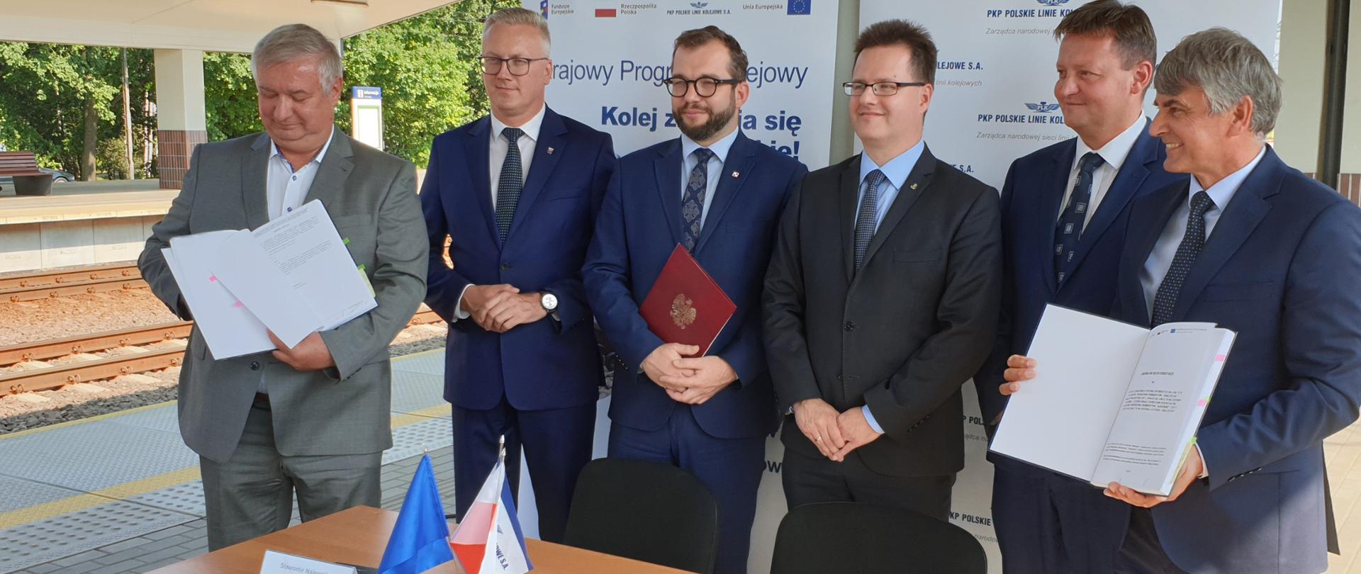 Szczęściu mężczyzn stoi przy stole, wśród nich minister Grzegorz Puda, dwóch z nich trzyma w ręku podpisane umowy. Stoją na peronie kolejowym. 