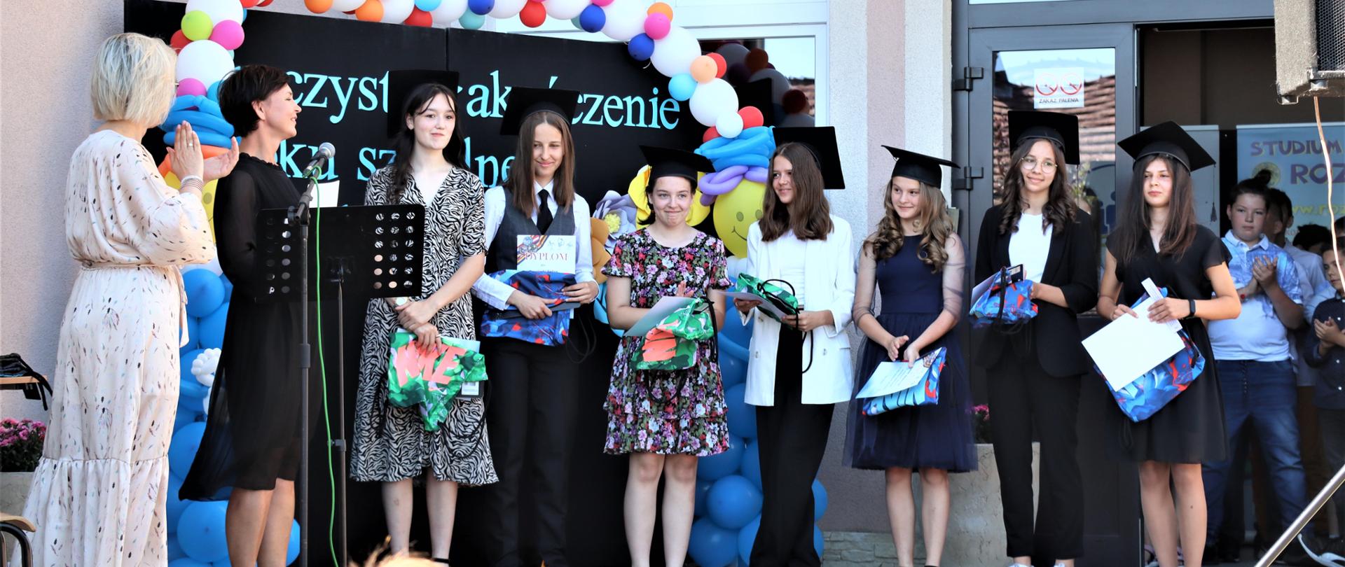 Uczniowie, ubrani w birety na głowach, którzy stoją na scenie. 