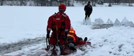 Strażacy ćwiczą na oblodzonym akwenie wodnym, widać wykuty przerębel i strażaków na desce lodowej. W tle drzewa oraz zabudowania. Obok przerębla ustawione na sztorc kry lodu.