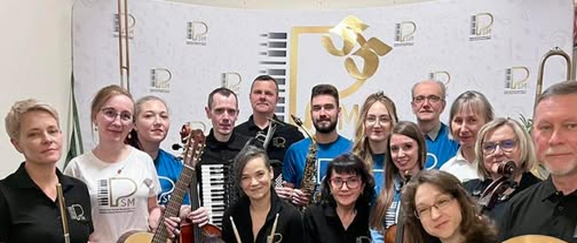 Kadra Państwowej Szkoły Muzycznej I stopnia w Lublińcu z instrumentami na tle ścianki z logo szkoły zachęcająca do nauki gry na instrumentach 