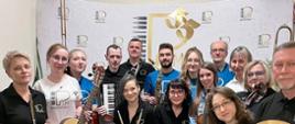 Kadra Państwowej Szkoły Muzycznej I stopnia w Lublińcu z instrumentami na tle ścianki z logo szkoły zachęcająca do nauki gry na instrumentach 