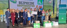 Scena: w centralnym miejscu, na całej długości sceny stoi piętnaście osób. W tle za nimi wyświetlany jest napis Bezpieczne Gospodarstwo Rolne. Przed nimi stoją nagrody. Dwóch mężczyzn w środku prezentuje zieloną statuetkę i voucher na 2000 zł. 