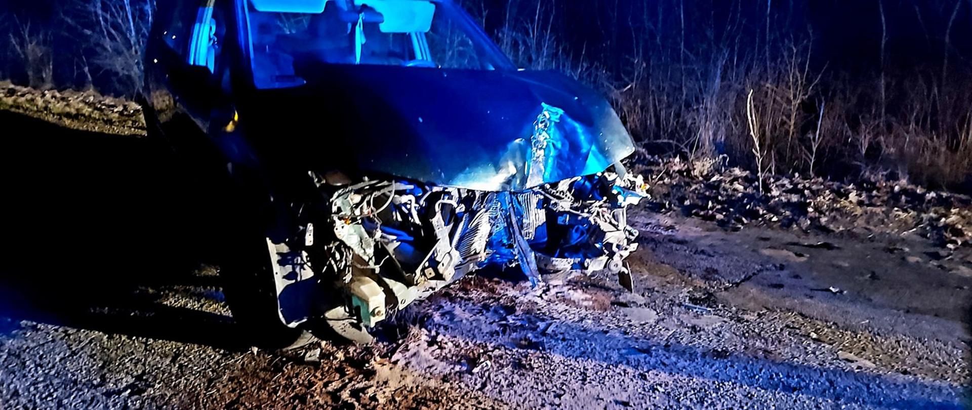 Zdjęcie przedstawia rozbite auto na drodze w porze nocnej. W tle widać drzewa.