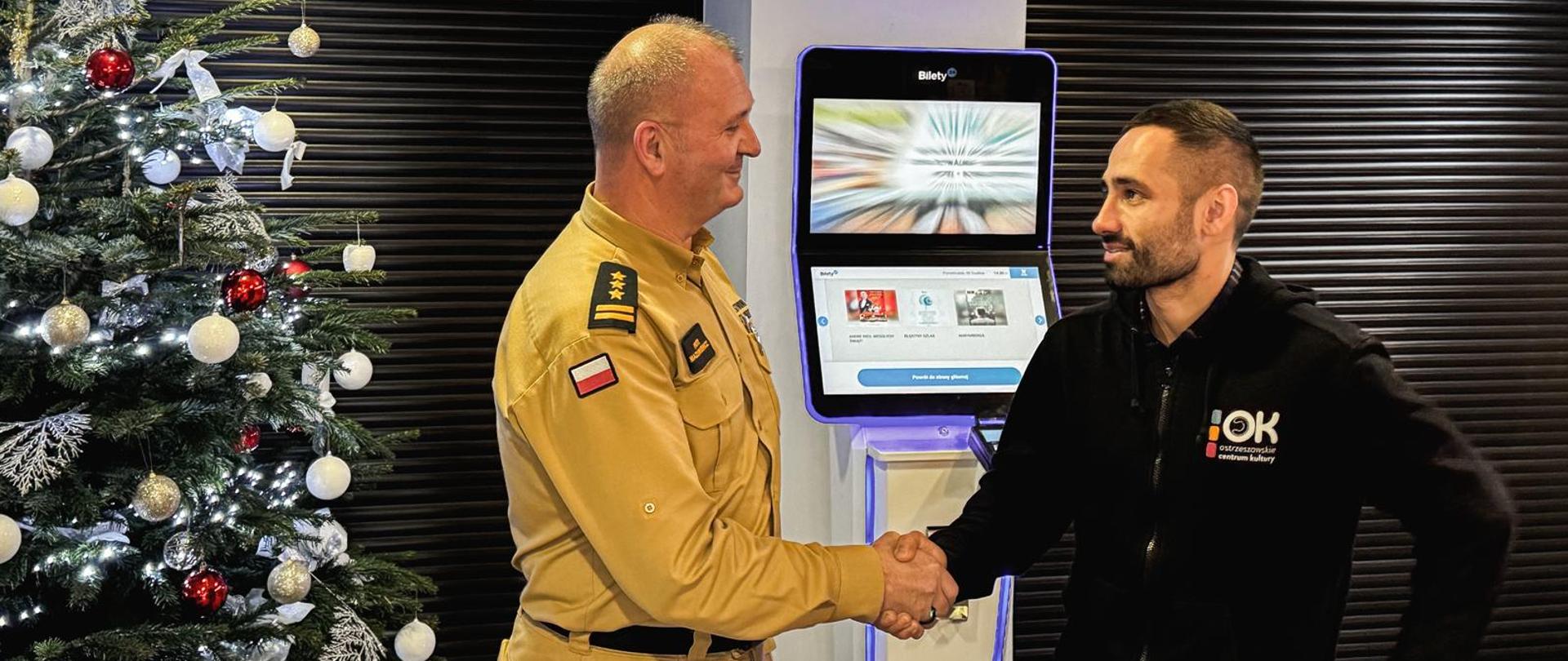 Komendant Powiatowy Państwowej Straży Pożarnej ściska dłoń dyrektorowi Centrum Kultury. W tle urządzenia elektroniczne, obok choinka. 