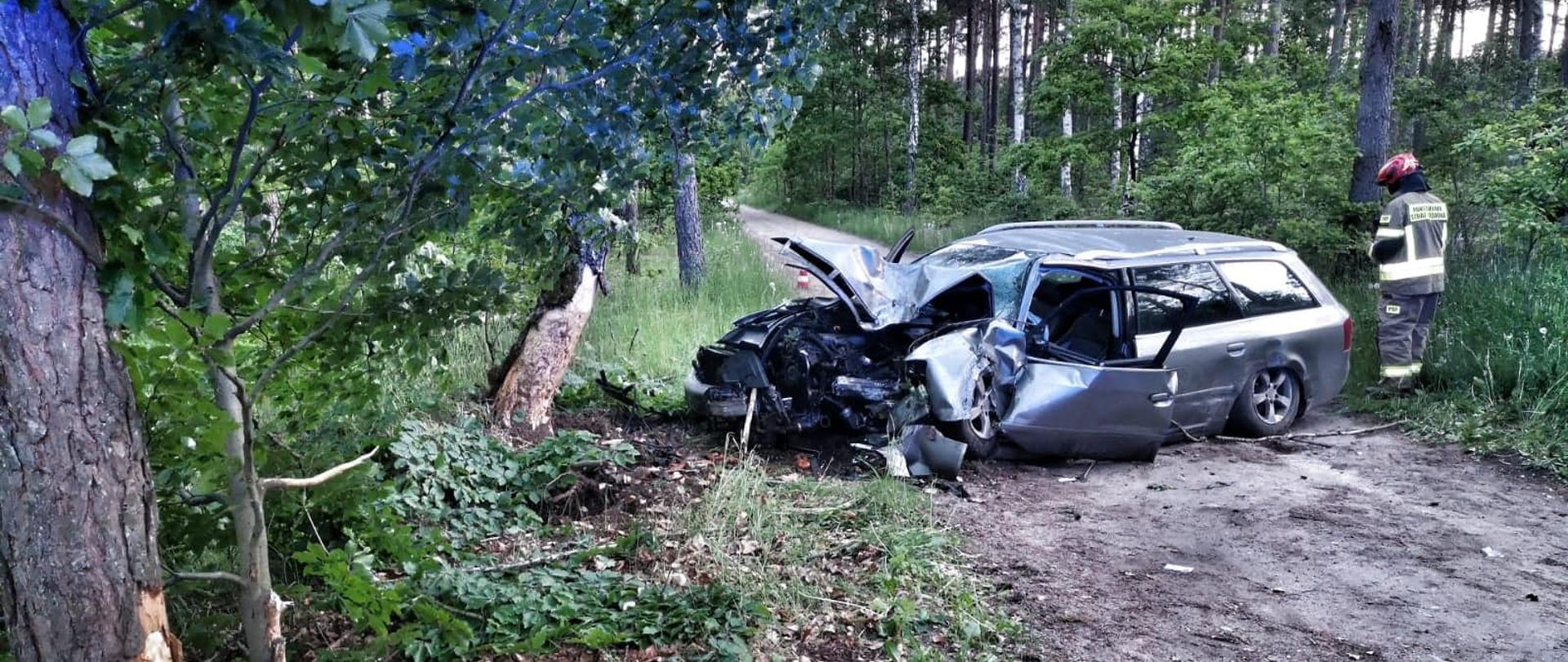 Na fotografii znajduje się rozbity pojazd, który znajduje się na środku dojazdu pożarowego w lesie. Pojazd ma rozbity przód oraz otwarte drzwi od strony kierowcy. Po prawej stronie fotografii znajduje się strażak. W tle znajdują się krzewy oraz drzewa.