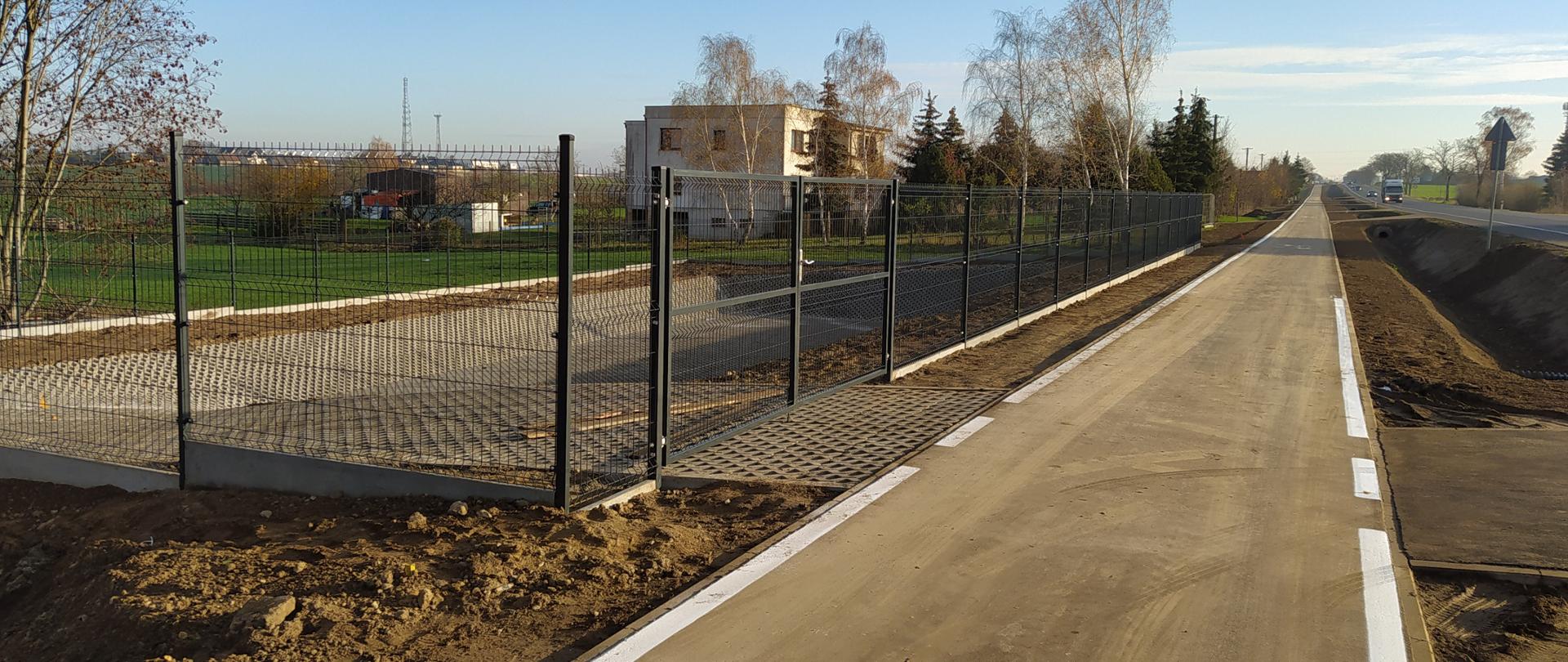 widok ścieżki rowerowej, po lewej stronie widoczny za płotem budynek mieszkalny, po lewej stronie droga krajowa, po której poruszają się pojazdy