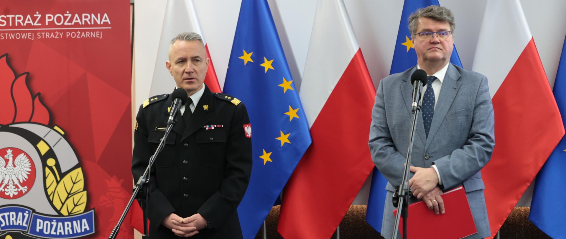 Strażak stoi przy mikrofonie w mundurze wyjściowym obok mężczyzna w garniturze trzyma czerwoną teczkę za nimi flagi Unii Europejskiej oraz Polski obok baner Komendy Głównej Państwowej Straży Pożarnej. 