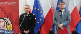 Strażak stoi przy mikrofonie w mundurze wyjściowym obok mężczyzna w garniturze trzyma czerwoną teczkę za nimi flagi Unii Europejskiej oraz Polski obok baner Komendy Głównej Państwowej Straży Pożarnej. 