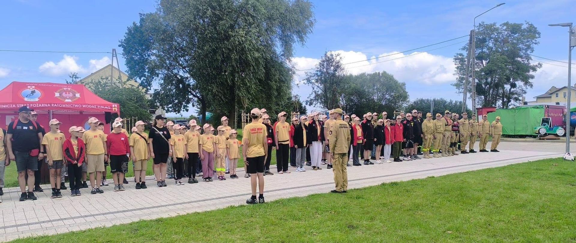 Na zdjęciu widać uczestników obozu MDP ustawionych w szyku podczas zbiórki. Młodzież oraz strażacy stoją w kolumnach, a prowadzący wydaje instrukcje przed rozpoczęciem zajęć.