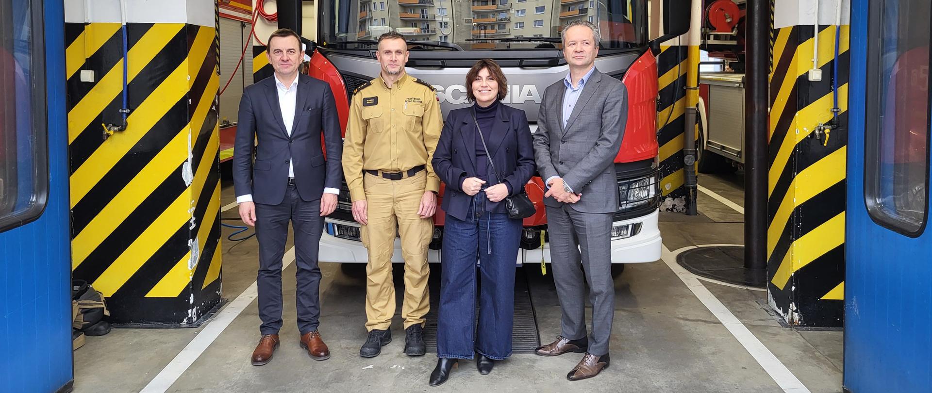 Na tle wozu strażackiego stoi komendant oraz zarząd Grupy Przemysłowej Baltic Sp.z o.o. w garażu PSP.