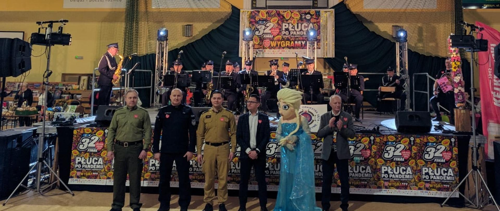 Na pierwszym planie widoczni przedstawiciele służb mundurowych (w tym p.o. Komendant Powiatowy PSP w Kętrzynie mł. bryg. Kamil Golon), starosta kętrzyński, maskotka – królewna oraz burmistrz miasta Kętrzyna. Za nimi na scenie gra Strażacka Orkiestra Dęta Ochotniczej Straży Pożarnej w Kętrzynie.