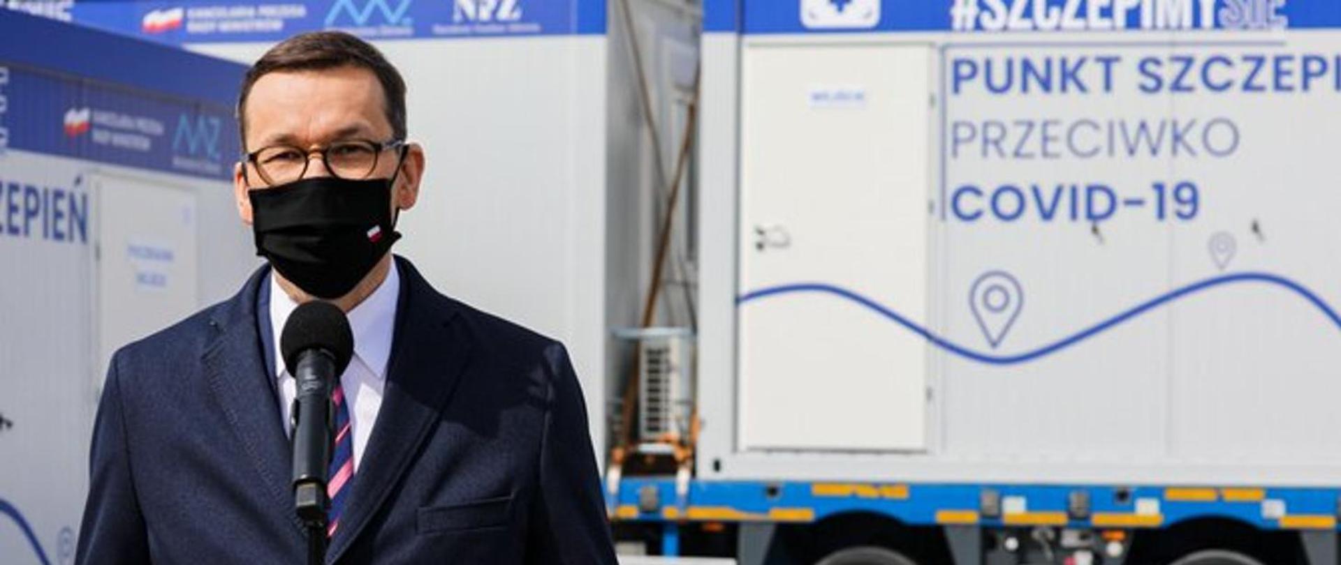 Na pierwszym planie Premier Polski - Mateusz Morawiecki - wyposażony w czarną maseczkę przed czarnym mikrofonem. W tel mobilne punkty szczepionek z niebieskim napisem na białym tle - punkt szczepień przeciwko -19