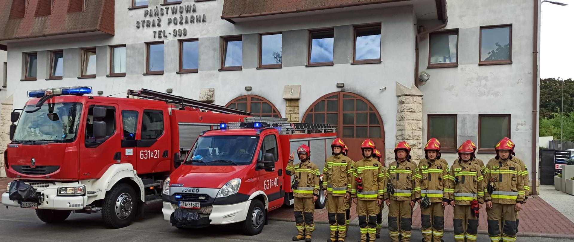 Strażacy w umundurowaniu stoją na baczność przed budynkiem jednostki straży pożarnej. Obok dwa samochody strażackie czerwone