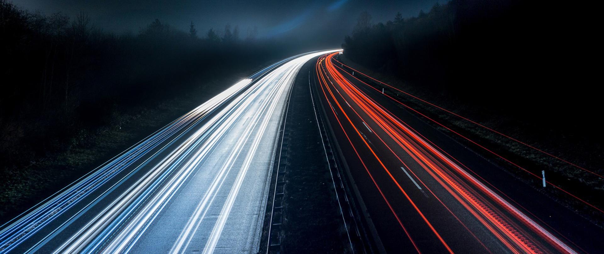 zdjęcie drogi nocą: światła przejeżdżających samochodów tworzą równoległe linie światła białego i czerwonego