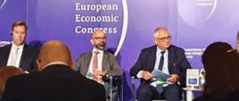 Wiceminister rozwoju i technologii Grzegorz Piechowiak siedzący z kartkami w dłoniach podczas panelu poświęconego inwestycjom zagranicznym na Europejskim Kongresie Gospodarczym w Katowicach, obok siedzą pozostali uczestnicy debaty, za ich plecami niebieski baner z logo EKG