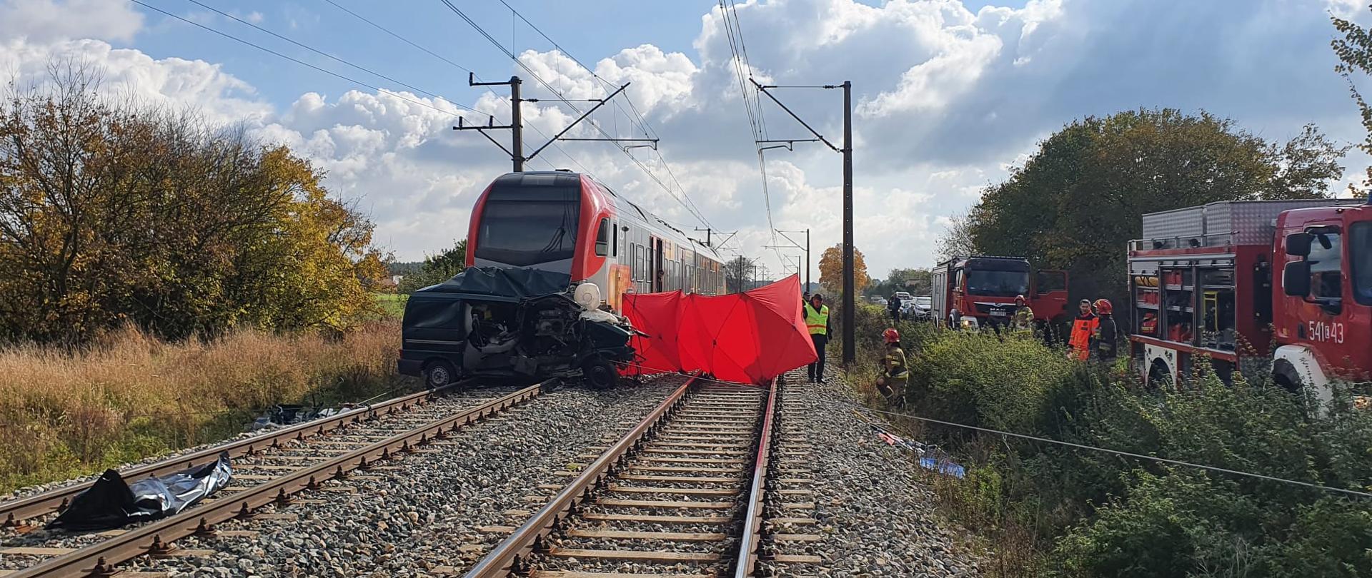 Na torach stoi pociąg. Przed pociągiem jest rozbity samochód. Z prawej strony pociągu znajduje się czerwony parawan. Obok torów stoją samochody strażackie. W tle drzewa i krzewy. 