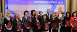 Zdjęcie zbiorowe, pod ścianą z pionowych drewnianych listewek z wbudowanym wielkim ekranem z napisem Powołanie Rady ds. Kobiet w Szkolnictwie Wyższym i Nauce stoi grupa kilkunastu kobiet trzymających czerwone teczki, pośrodku grupy minister Kulasek i wiceminister Zioło-Pużuk.