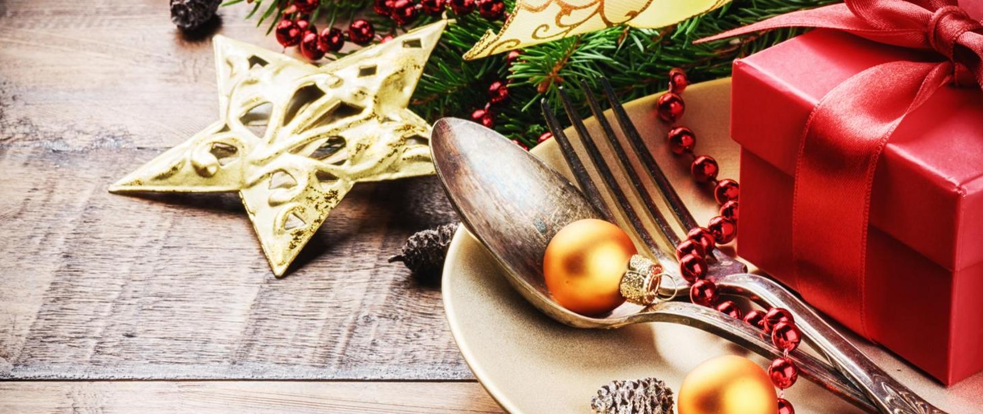 Na zdjęciu widoczny talerz, sztućce, na talerzu czerwone pudełko i dwie złote bombki a obok talerza ozdoba świąteczna w postaci świerku, gwiazdy i szyszek.