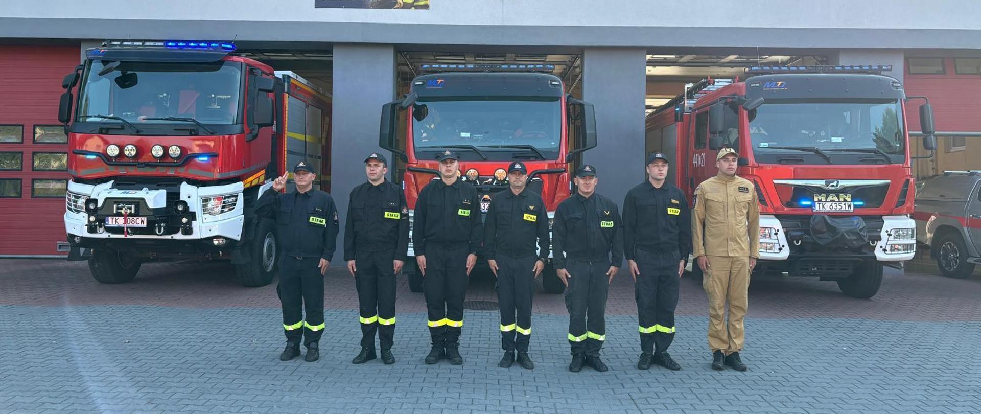 Grupa strażaków w ubraniach koszarowych przed samochodami pożarniczymi z włączonymi sygnałami świetlnymi niebieskimi na placu zewnętrznym KPPSP W Opatowie