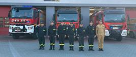 Grupa strażaków w ubraniach koszarowych przed samochodami pożarniczymi z włączonymi sygnałami świetlnymi niebieskimi na placu zewnętrznym KPPSP W Opatowie