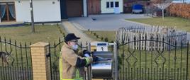 strażak OSP wrzuca ulotkę na temat szczepienia do skrzynki pocztowej, w tle budynek z podjazdem.
