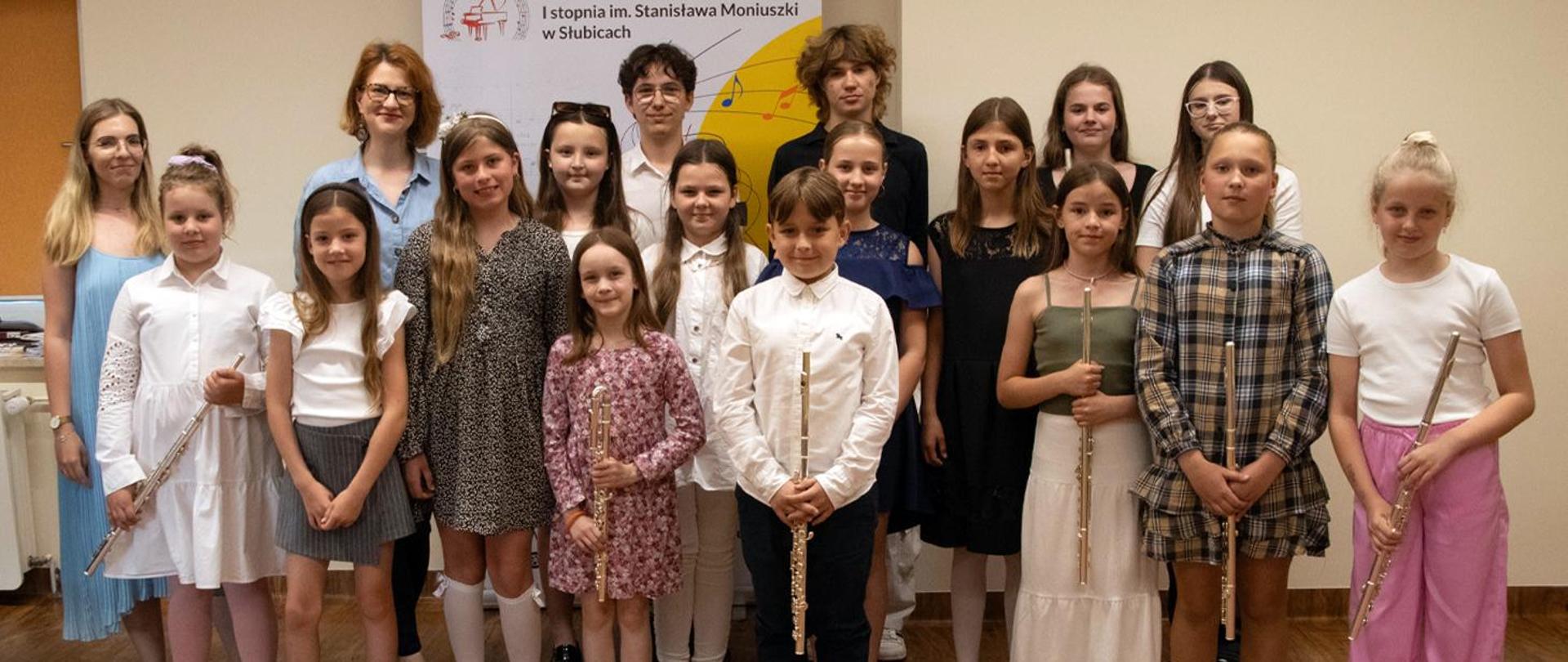 Na zdjęciu kilkunastoosobowa grupa uczniów i nauczycieli stoi a w auli szkolnej. Niektóre z dzieci w rękach trzymają flety. Za nimi baner szkoły.