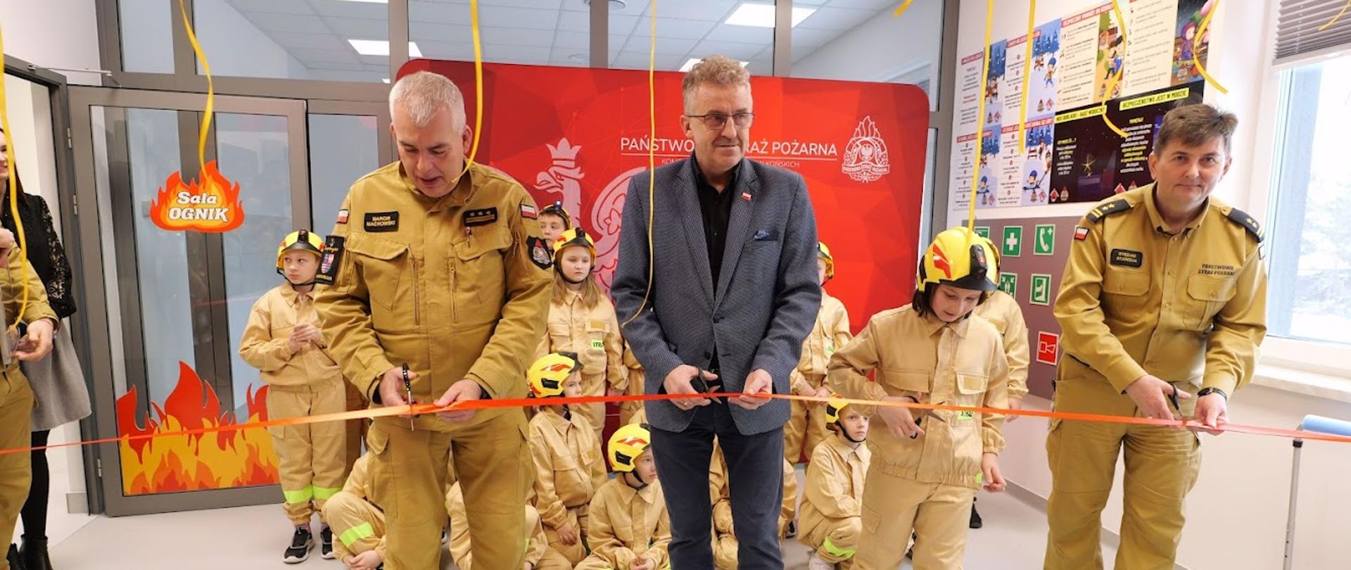 Trzech dorosłych mężczyzn stoi na pierwszym planie i przecina czerwoną wstęgę podczas uroczystego otwarcia sali edukacyjnej w Komendzie Państwowej Straży Pożarnej. Dwóch z nich ubranych jest w piaskowe mundury strażackie PSP, a jeden w środkowej części kadru ma na sobie granatową marynarkę i koszulę. Obok nich znajduje się dziecko w stroju małego strażaka, również trzymające nożyczki. W tle widoczna jest grupa dzieci ubranych w beżowe stroje strażackie i żółte hełmy ochronne, siedzących i stojących przed czerwoną planszą z napisem „Państwowa Straż Pożarna” oraz godłem Polski. Nad uczestnikami wiszą dekoracyjne, żółte wstążki. Po lewej stronie widoczne są drzwi z napisem „Sala Ognik” i motywem płomieni. Scena przedstawia oficjalne otwarcie przestrzeni edukacyjnej z udziałem strażaków i dzieci.