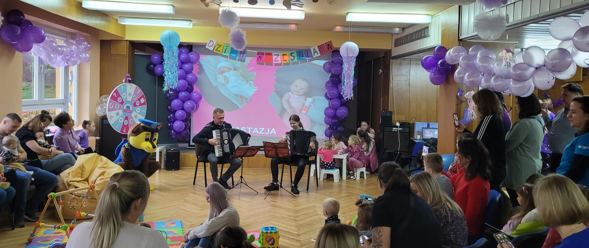 Nauczyciel wraz z uczennicą gra na akordeonie. Wokół siedzi publiczność. Salę dekorują kolorowe balony oraz wiszący napis "dzień wcześniaka"
