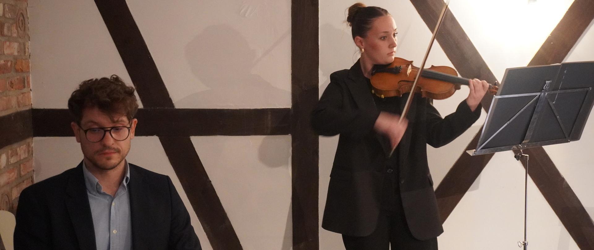 Na zdjęciu po lewej stronie mężczyzna w błękitnej koszuli i czarnej marynarce, w ciemnych kręconych włosach oraz okularach, który gra na pianinie. Po prawej stronie dziewczyna w czarnych spodniach i czarnej marynarce, która gra na skrzypcach. Przed nią pulpit z nutami. Zdjęcie wykonane zostało w kameralnym wnętrzu podczas uroczystości podpisania porozumienia między gminą Stara Kiszewa a Gminą Lahntal.