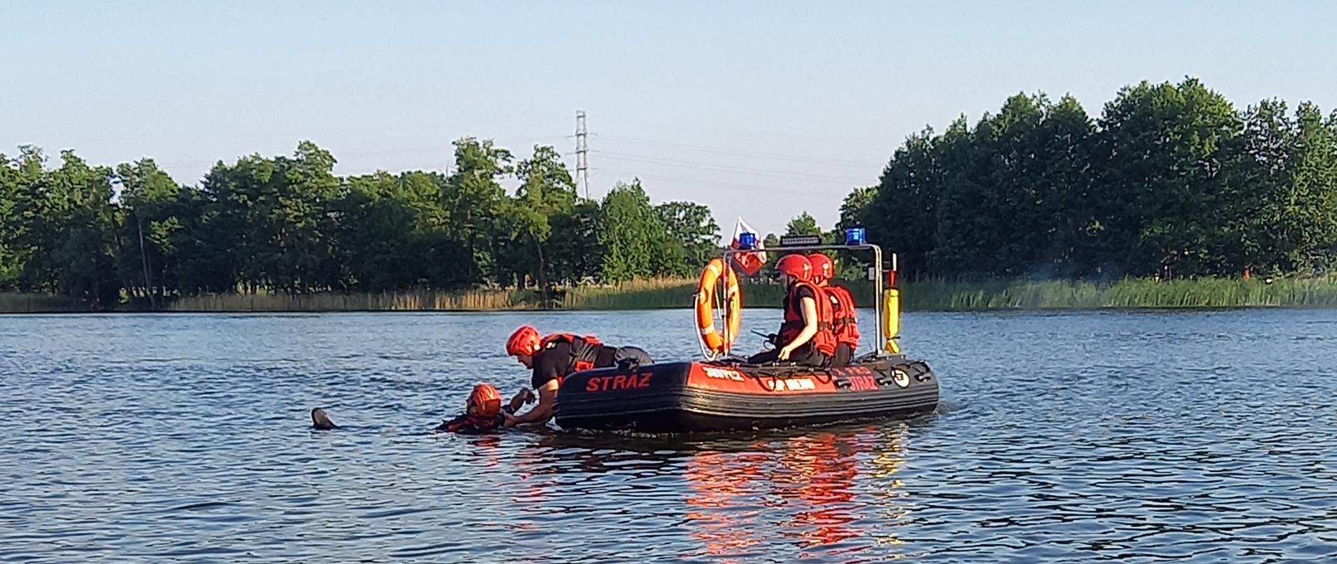 Trzech strażaków zabezpieczonych w kamizelki ratownicze, z łodzi pontonowej podejmuje osobę poszkodowaną z wody.