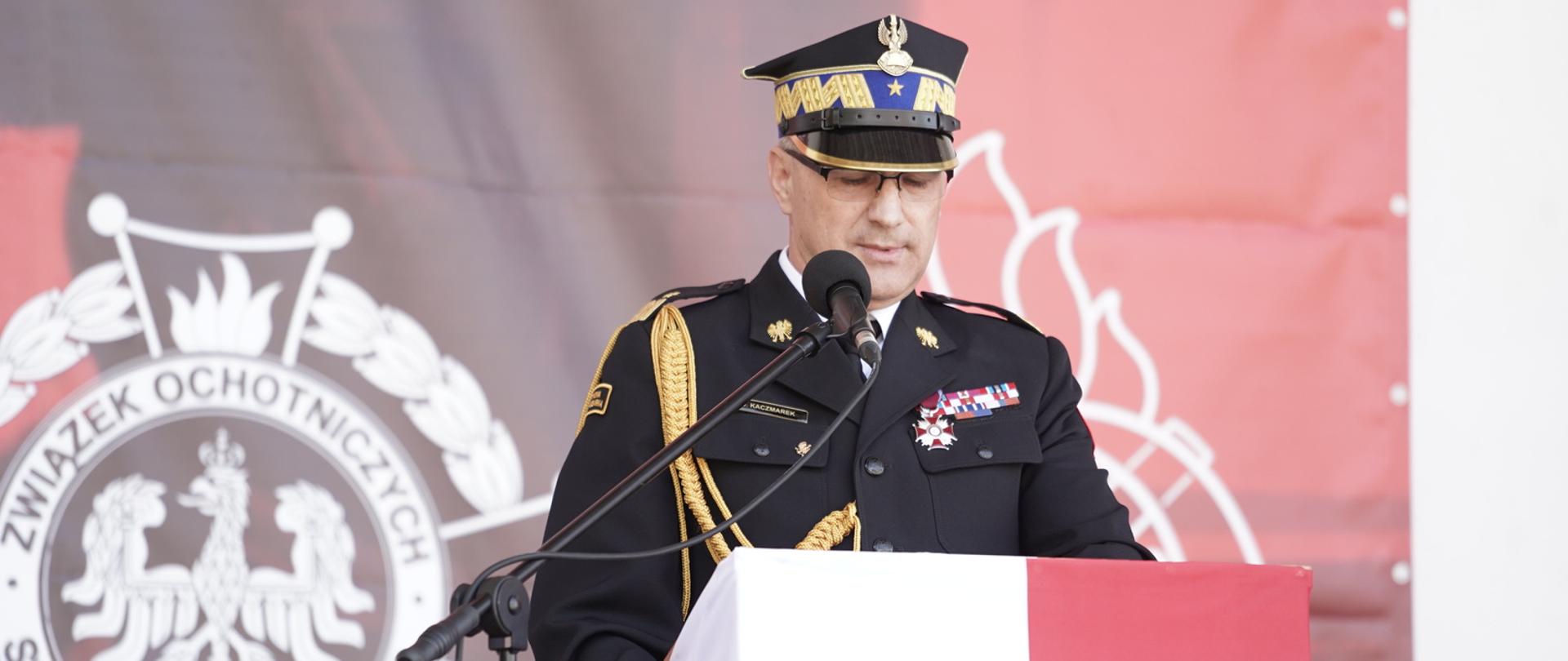 Komendant Wojewódzki PSP przy mównicy. Ubrany jest na galowo, w czapce. Mównica obleczona flagą biało-czerwoną. W tle baner okolicznościowy święta.