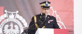 Komendant Wojewódzki PSP przy mównicy. Ubrany jest na galowo, w czapce. Mównica obleczona flagą biało-czerwoną. W tle baner okolicznościowy święta.