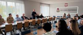 Spotkanie przedstawicieli samorządów i strażaków w sali konferencyjnej KP PSP w Radomsku. Funkcjonariusz w ciemnym mundurze przemawia do uczestników siedzących przy stołach. W tle widoczne są godło Polski oraz emblemat Państwowej Straży Pożarnej.