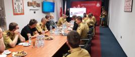 Zdjęcie przedstawia strażakom siedzących przy stole w sali konferencyjnej. Zastępca komendanta miejskiego Państwowej Straży Pożarnej omawia prezentację dotyczącą łączności.