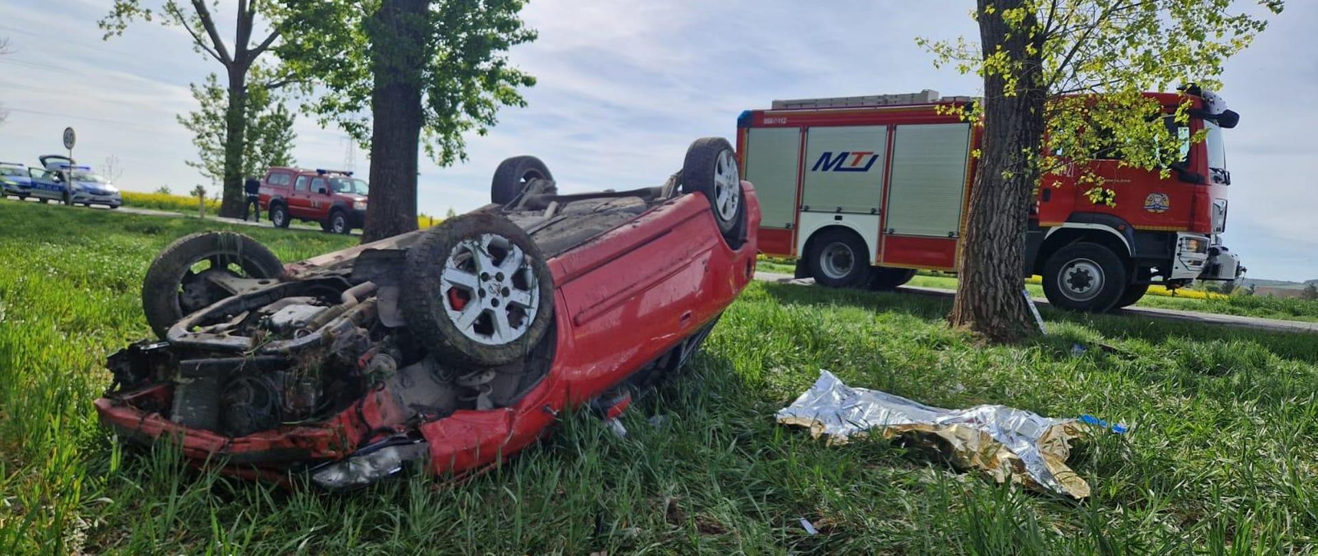 Samochód osobowy leży na dachu w polu. W tle samochody pożarnicze i radiowóz policyjny