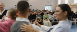 Na pierwszym planie para tańcząca w stroju ludowym, w tle młodzież szkolna oglądająca występ.