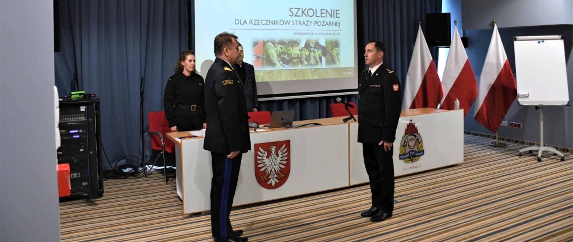 Na zdjęciu widocznych dwóch oficerów PSP stojących do siebie frontem. Funkcjonariusze w mundurach wyjściowych. Oficer po prawej stronie składa meldunek stojącemu po lewej stronie komendantowi wojewódzkiemu PSP. W tle widać stół konferencyjny z herbem województwa i logiem PSP oraz ekran, na którym wyświetlony jest pierwszy slajd prezentacji z tytułem "Szkolenie dla rzeczników straży Pożarnej". W rogu stali stoją trzy biało-czerwone flagi.