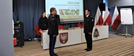 Na zdjęciu widocznych dwóch oficerów PSP stojących do siebie frontem. Funkcjonariusze w mundurach wyjściowych. Oficer po prawej stronie składa meldunek stojącemu po lewej stronie komendantowi wojewódzkiemu PSP. W tle widać stół konferencyjny z herbem województwa i logiem PSP oraz ekran, na którym wyświetlony jest pierwszy slajd prezentacji z tytułem "Szkolenie dla rzeczników straży Pożarnej". W rogu stali stoją trzy biało-czerwone flagi.