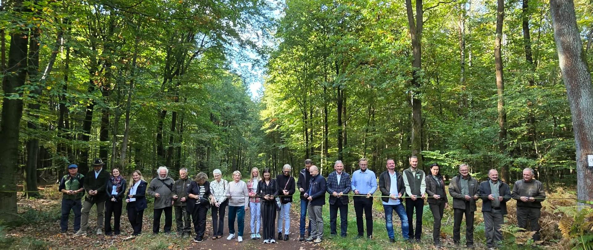Las. Liczna grupa osób stoi w lesie przed rozciągniętą wstęgą. Kilka z nich przecina szarfę.