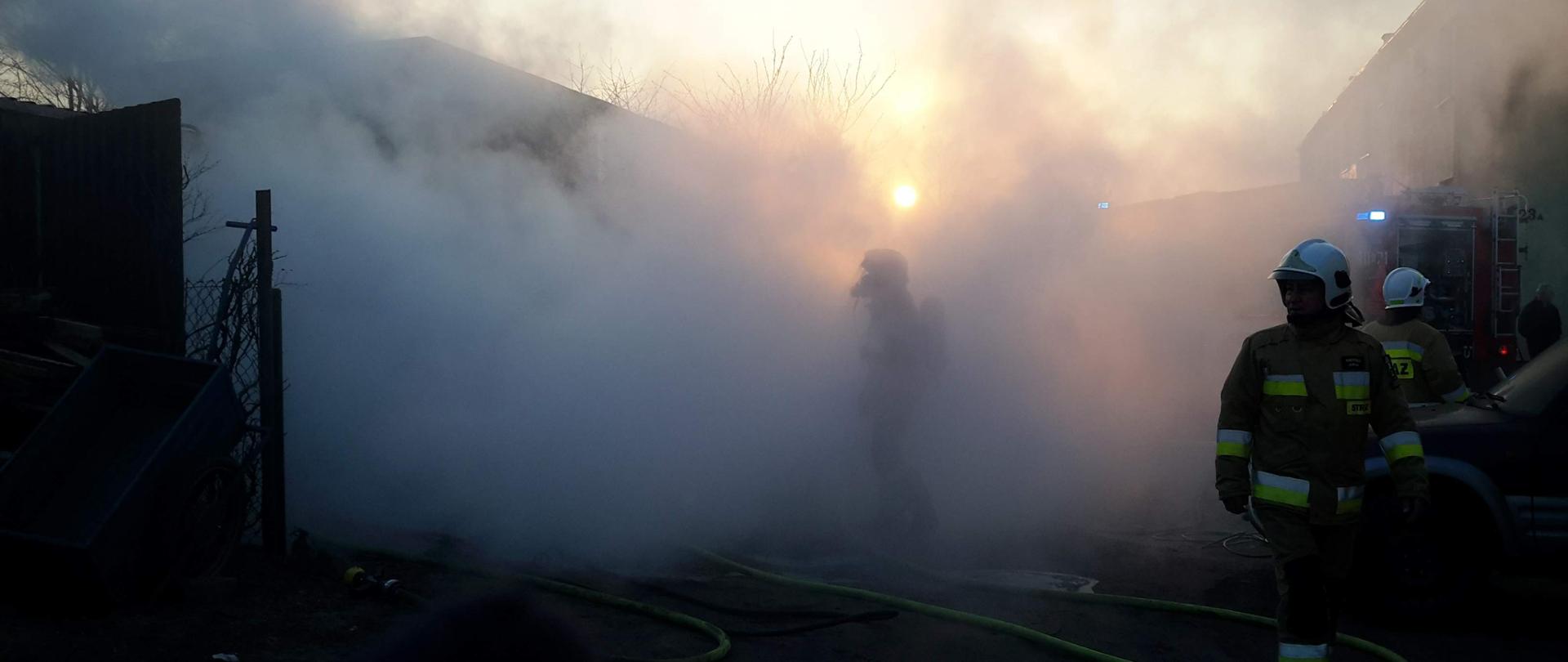 Strażacy w aparatach oddechowych w czasie gaszenia garażu. Duże zadymienie. W tle wschód słońca.