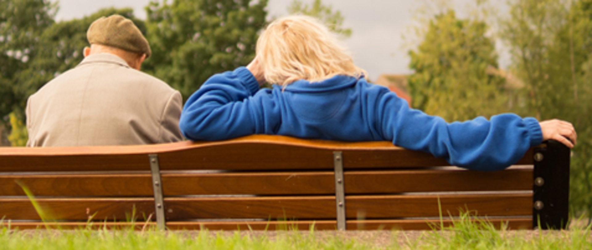 Na zdjęciu przedstawiona jest para seniorów siedząca tyłem na drewnianej ławce. Po lewej stronie siedzi mężczyzna w beżowym płaszczu i kaszkiecie w kolorze khaki. Po prawej stronie siedzi kobieta w długich blond włosach oraz ciemnoniebieskiej polarowej bluzie. W tle widać drzewa oraz zarośla co sugeruje, że para znajduje się w parku.