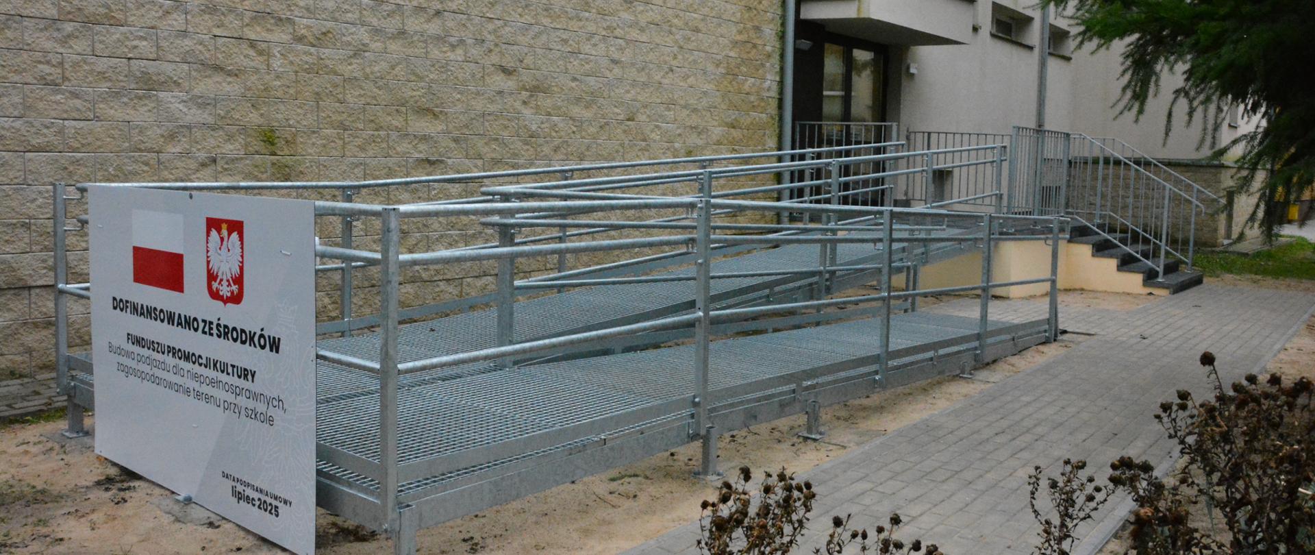 Metalowa rampa , schody oraz wejście do budynku , na rampie tablica informująca o dofinansowaniu z Funduszy Promocji Kultury, chodnik z brukowej kostki