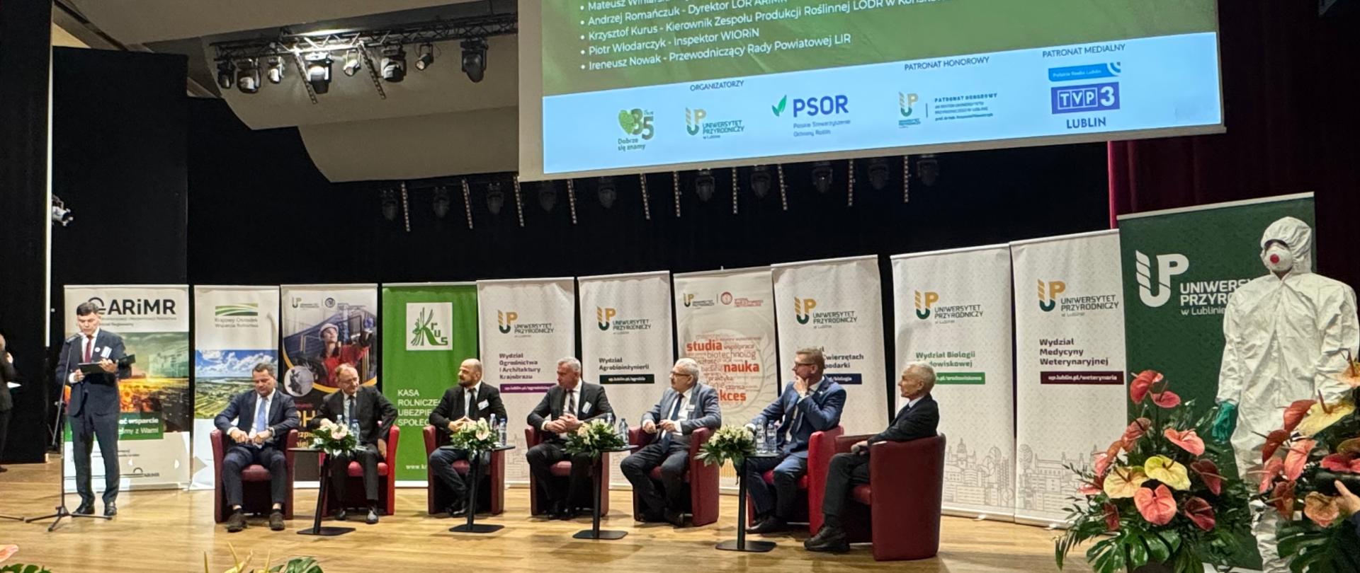 Panel dyskusyjny o bezpiecznym stosowaniu środków ochrony roślin na scenie; kilku ekspertów siedzi w fotelach pod dużym ekranem z tytułem wydarzenia, obok stoi osoba w stroju ochronnym
