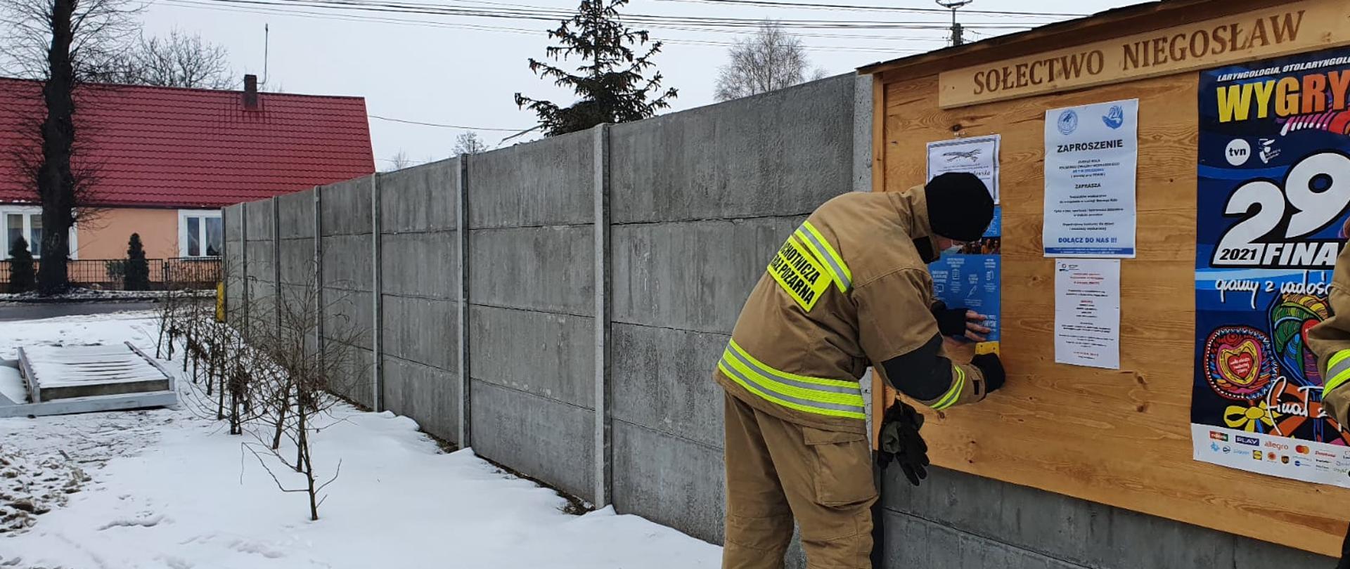 Fotografia przedstawia strażaka ochotnika w umundurowaniu specjalnym umieszczającego plakat informacyjny dotyczący szczepień na tablicy ogłoszeń sołectwa Niegosław. Tablica ogłoszeń wisi na betonowym ogrodzeniu, na ziemi leży śnieg, w tle po lewej stronie widoczny budynek mieszkalny.