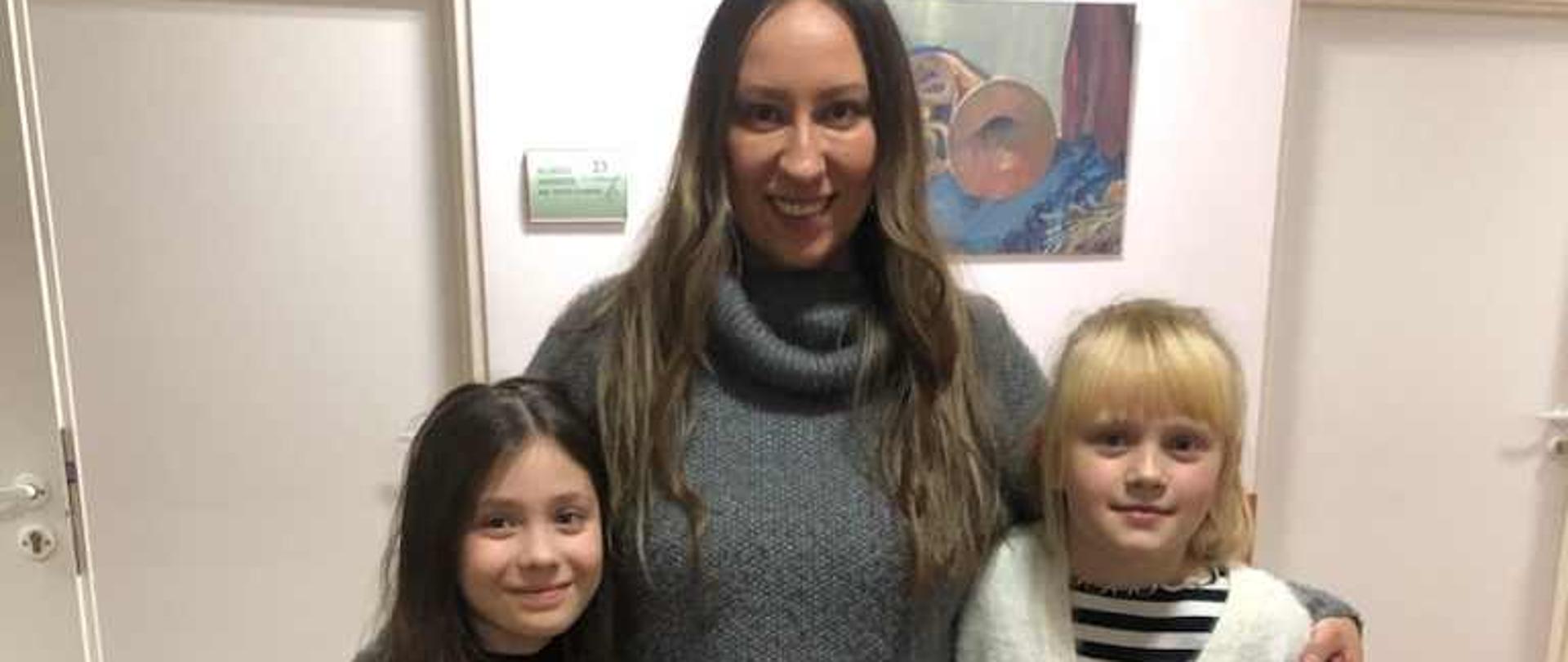 Zdjęcie przedstawia dwie uczennice po obu stronach pani która obejmuje je ramionami. W tle korytarz i i drzwi do sal lekcyjnych.
