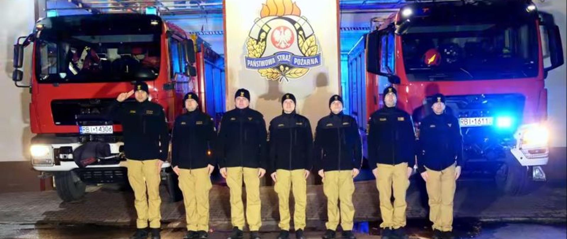 Na zdjęciu widać strażaków ustrzyckiej komendy którzy stoją na baczność za nimi samochody z włączonymi sygnałami świetlnymi. 