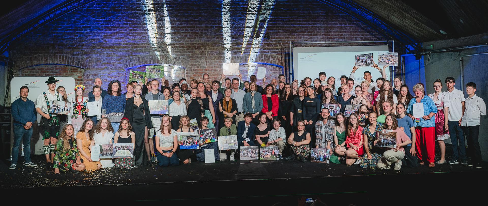 Widok na salę, pod półokrągłym sklepieniem stoi duża grupa kolorowo ubranej młodzieży.